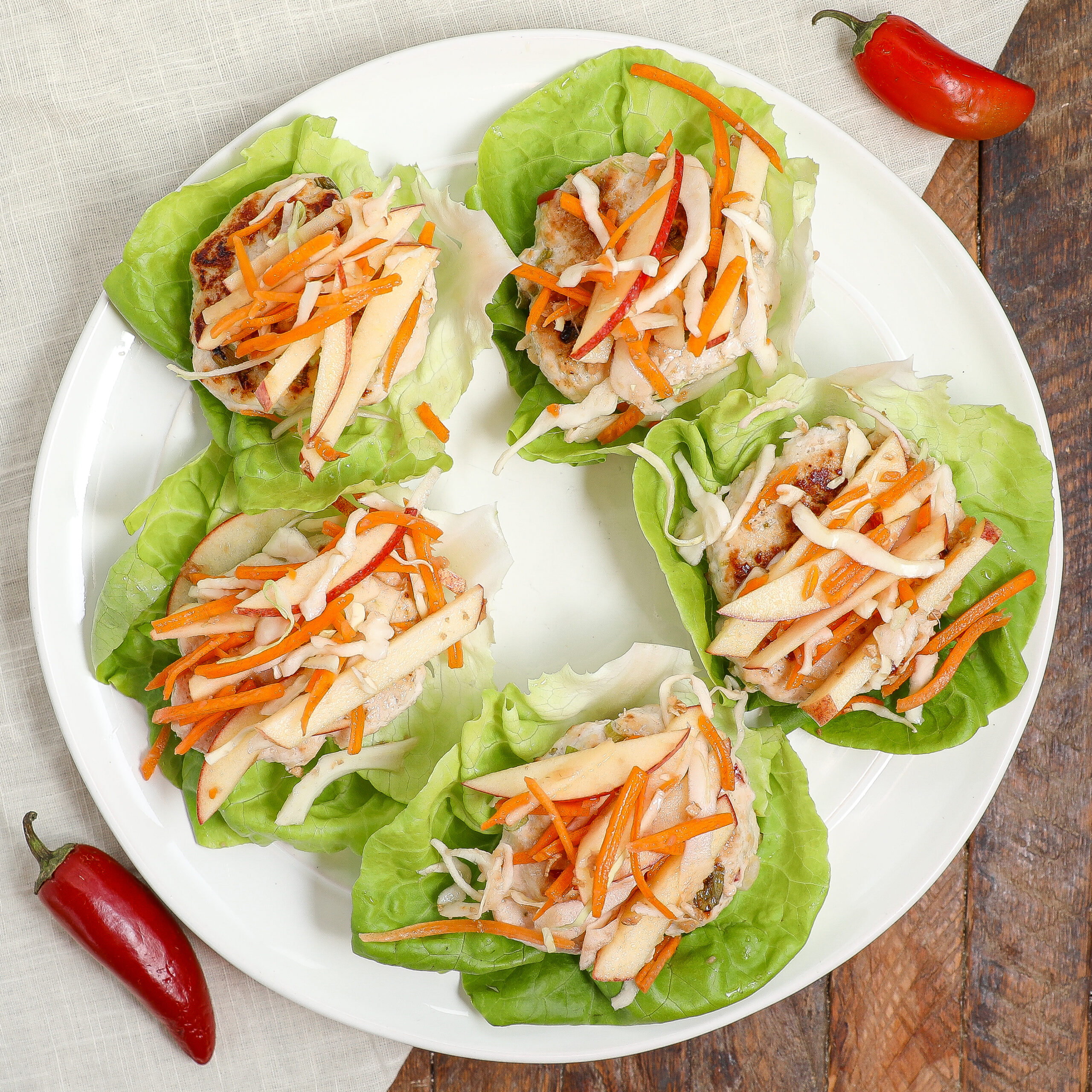 Five lettuce wraps filled with chicken patties, shredded carrots, cabbage, and apple slices are arranged on a white plate. Inspired by Sunbasket flavors, the dish sits on a wooden surface with two red chili peppers nearby.