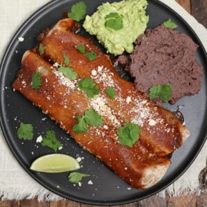 A black plate with two birria enchiladas topped with red sauce, cotija cheese, and cilantro, served with a scoop of guacamole, refried black beans, and a lime wedge on a light cloth napkin.