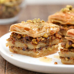 Close-up of delicious baklava on a white plate, highlighting its flaky layers of pastry, chopped nuts, and syrup, with a sprinkle of crushed pistachios on top.