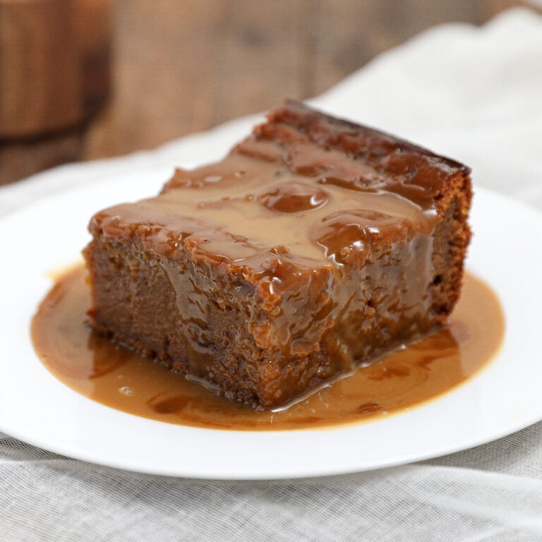 A slice of sticky toffee gingerbread pudding cake is served on a white plate, drenched in rich caramel sauce, with a light fabric and wooden surface in the background.