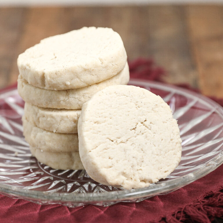 A stack of round, pale shortbread cookies sits on a clear glass plate, with one classic shortbread cookie resting in front. The plate is on a red cloth atop a wooden surface.