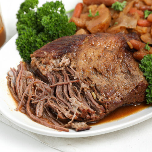 Close-up of oven braised brisket on a plate with gravy, carrots, potatoes, and parsley.