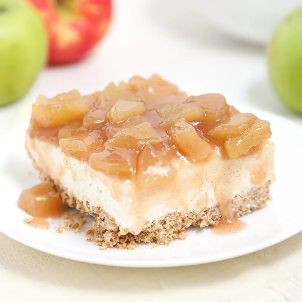 A slice of dessert with a crumbly base, creamy middle layer, and a topping of glazed apple chunks on a white plate. Whole apples are blurred in the background.