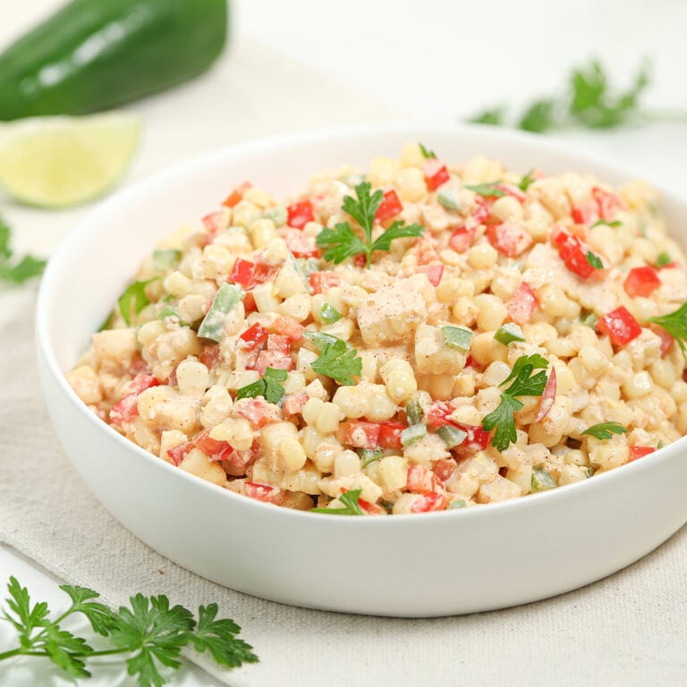 A white bowl filled with creamy corn salad, mixed with chopped red peppers, green herbs, and garnished with fresh parsley. A lime wedge and jalapeño are in the background on a light surface.