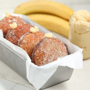 A metal loaf pan lined with parchment paper holds sugar-coated doughnuts filled with cream. In the background, two whole bananas and a jar of creamy filling are visible.