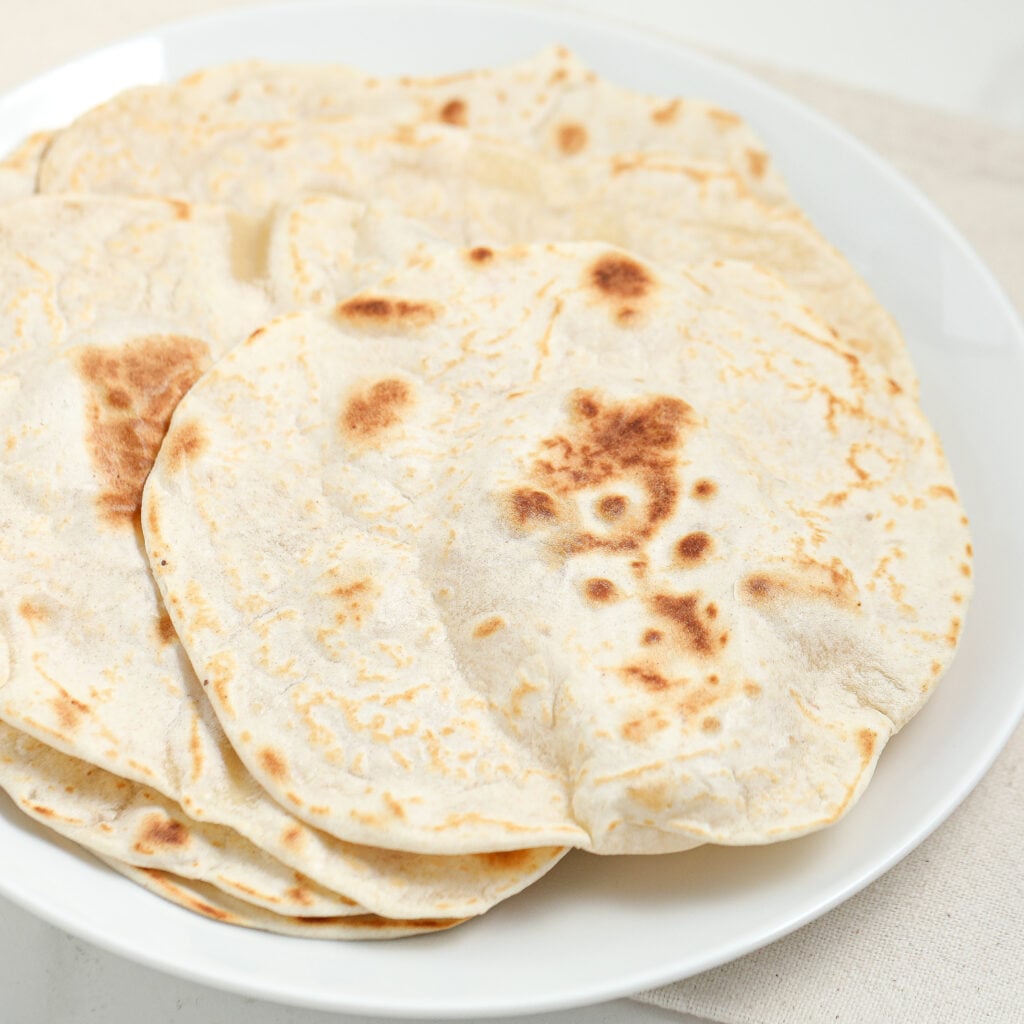 A white plate filled with several round, lightly browned flatbreads stacked on top of each other. The flatbreads have a soft, slightly puffy texture with golden spots.