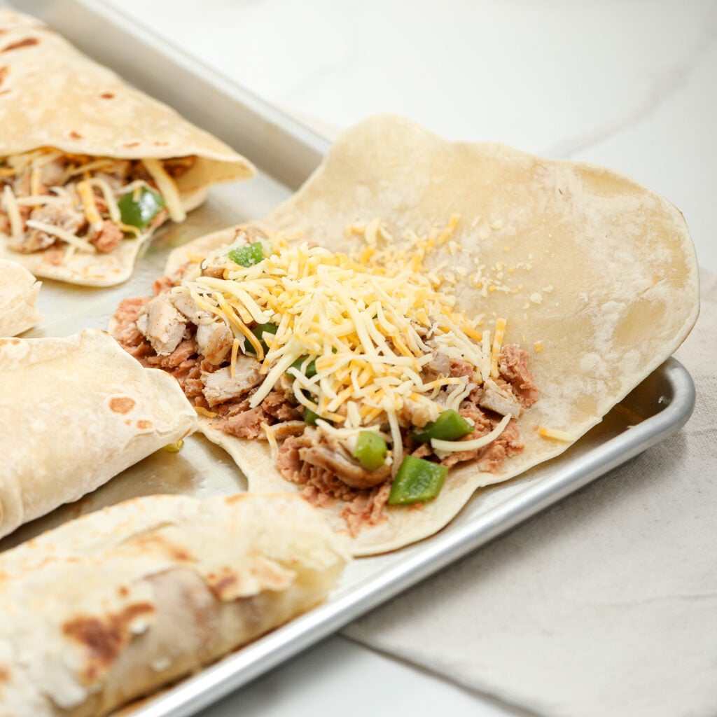 An open flour tortilla on a metal tray, filled with shredded chicken, refried beans, chopped green bell peppers, and shredded cheese, with two wrapped burritos nearby.