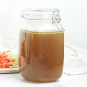 A glass jar filled with brown homemade broth sits on a white surface, with chopped carrots and celery on a cutting board in the blurred background.