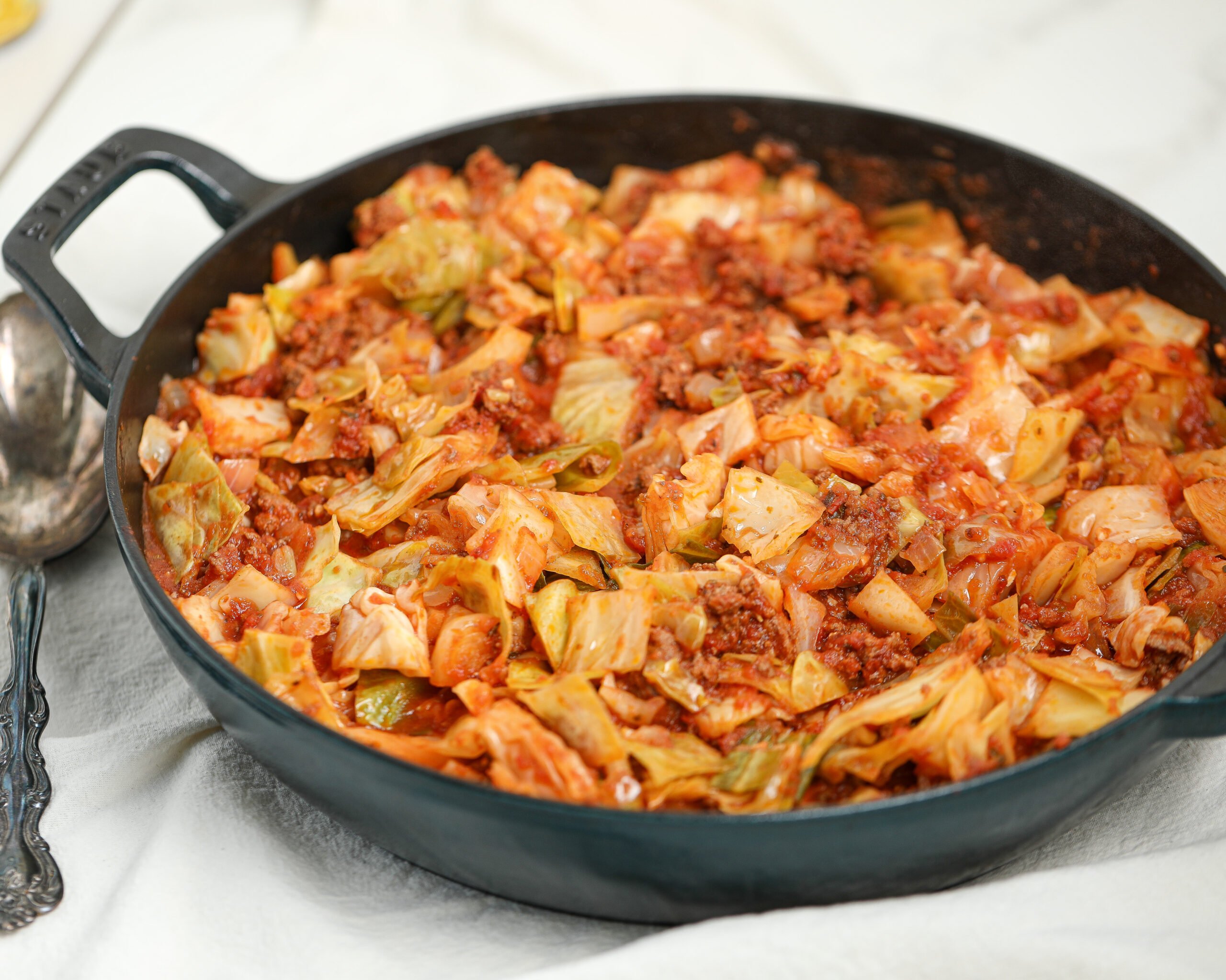 A skillet filled with old-fashioned Amish cabbage casserole, featuring chunks of cabbage, ground beef, and tomato sauce, sitting on a white cloth.