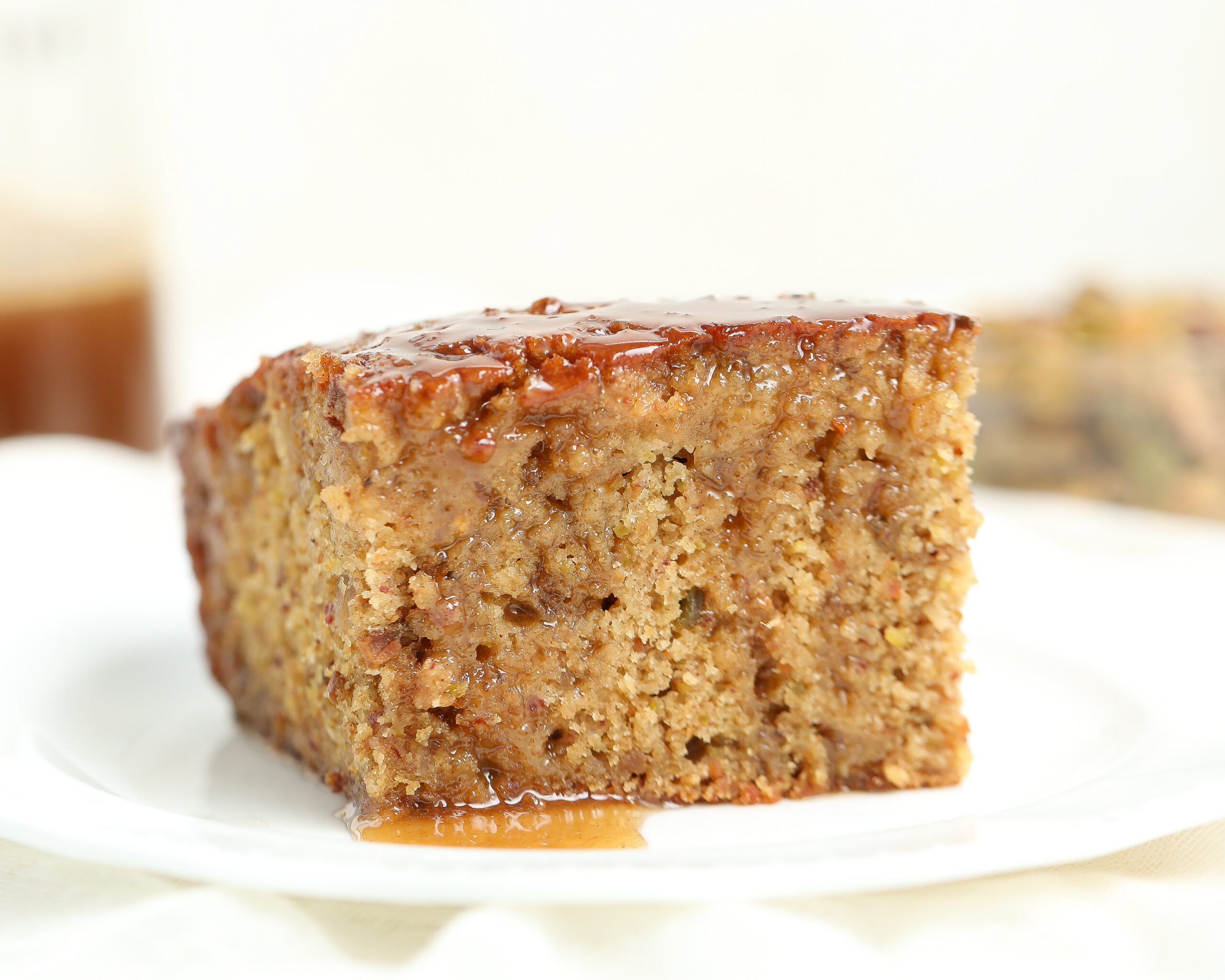 A close-up slice of Baklava Sticky Pudding Cake showing its dense, moist texture with visible date and pistachio specks, topped with glossy honey sauce dripping slightly down the sides.