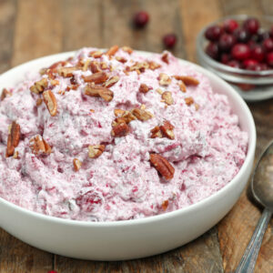 A white bowl filled with creamy pink cranberry fluff salad topped with pecans, on a wooden surface. A spoon and small bowl of fresh cranberries are visible in the background.