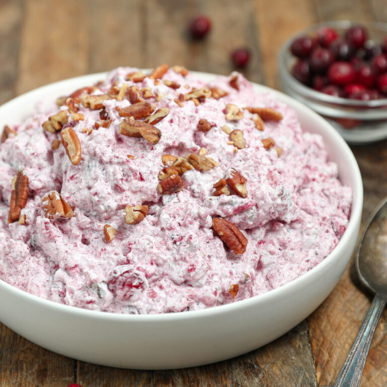 A white bowl filled with creamy pink cranberry fluff salad topped with pecans, on a wooden surface. A spoon and small bowl of fresh cranberries are visible in the background.