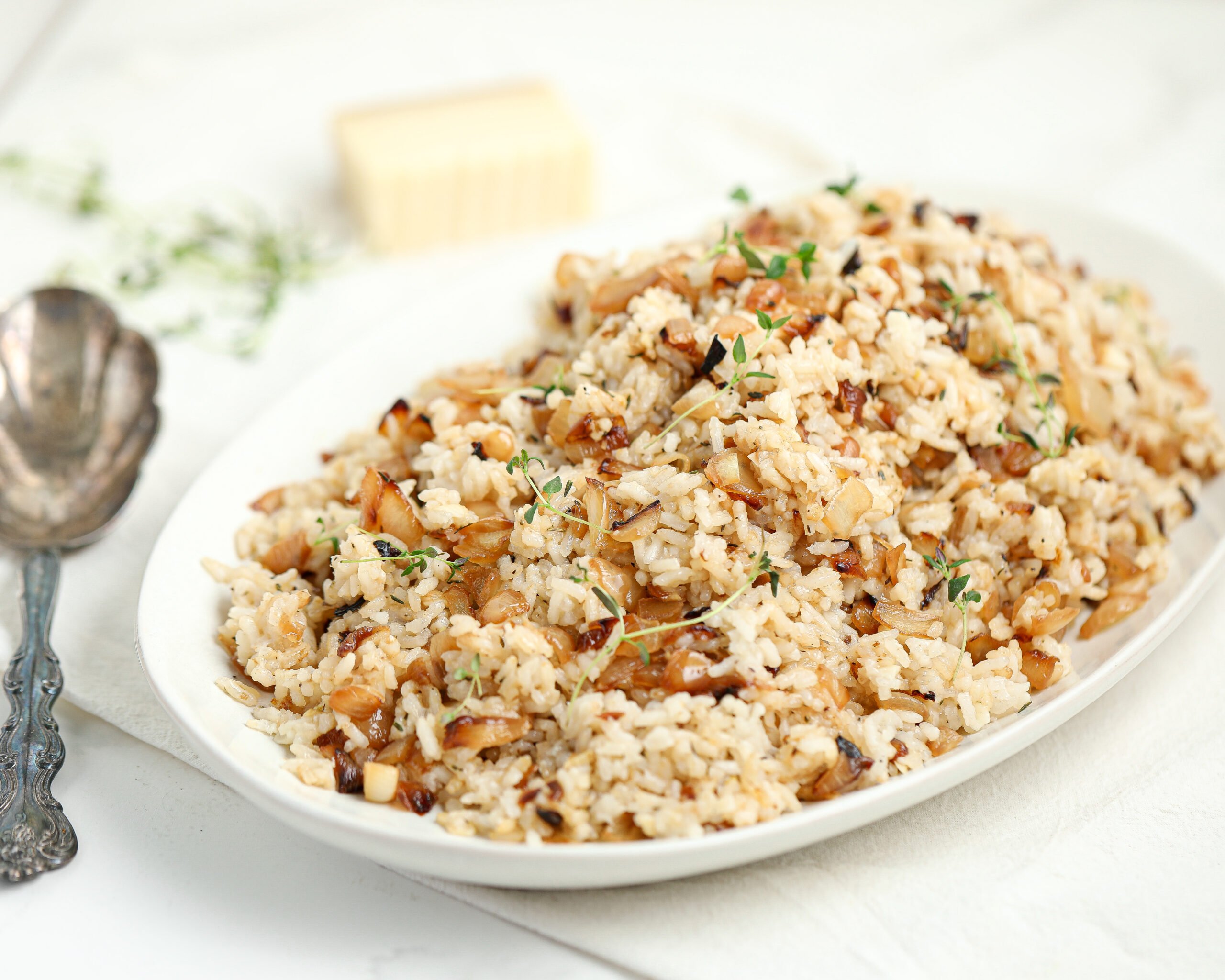 A white serving platter filled with fluffy rice mixed with golden caramelized onions and sprinkled with fresh thyme, sitting on a light tabletop with a block of cheese and vintage silverware nearby.
