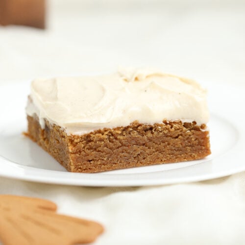 A square slice of gingerbread brownie topped with espresso cream cheese frosting on a white plate, with a blurred gingerbread cookie in the foreground.