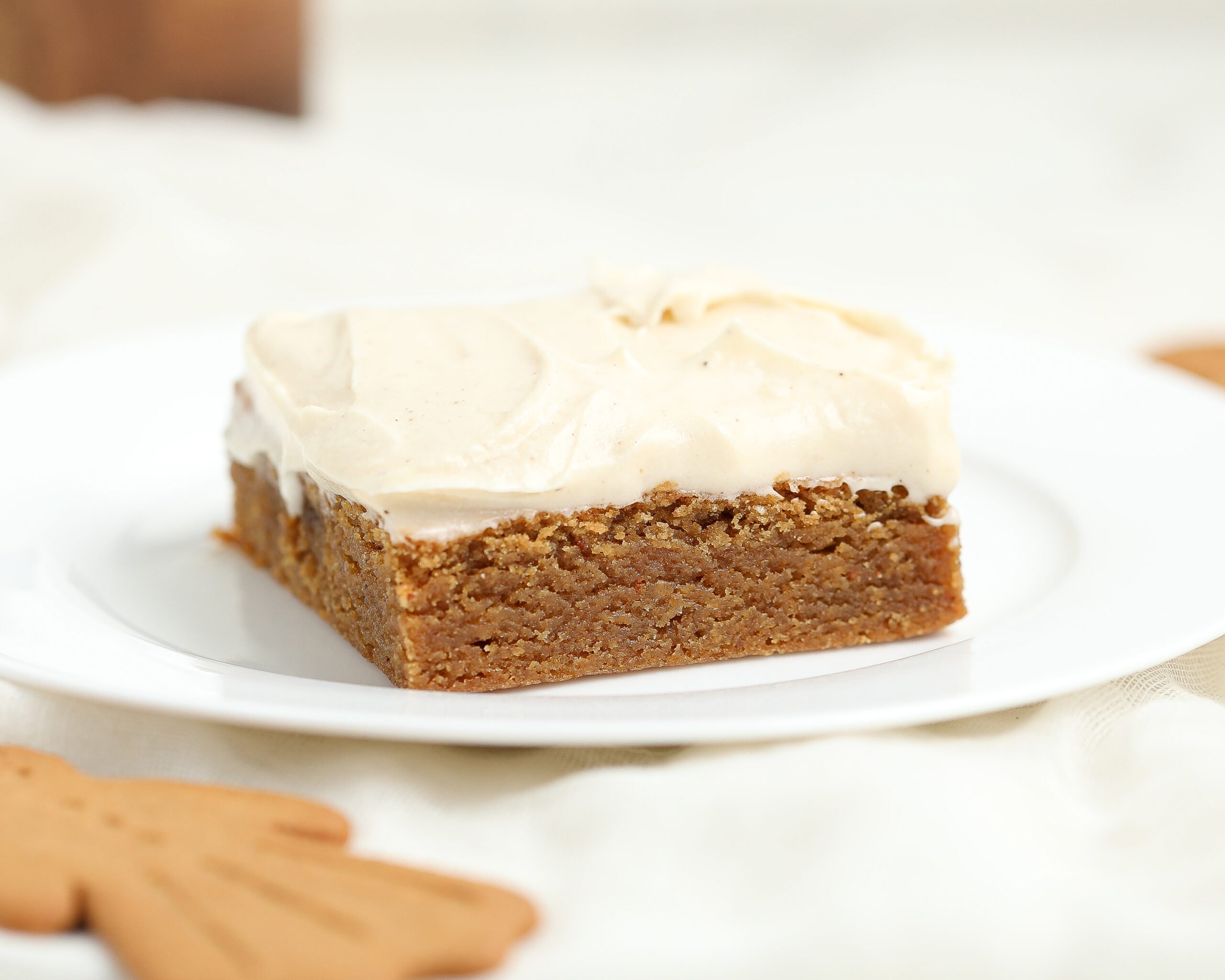 A square slice of gingerbread brownie topped with espresso cream cheese frosting on a white plate, with a blurred gingerbread cookie in the foreground.