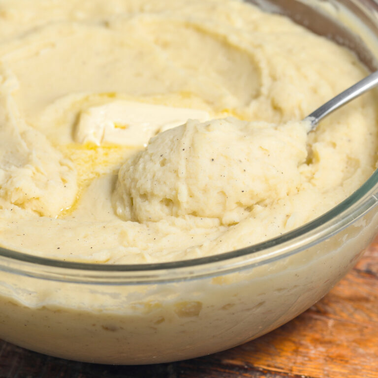 A glass bowl filled with creamy mashed potatoes, topped with a pat of melting butter. A metal spoon is scooping some of the mashed potatoes. The bowl is on a wooden surface.