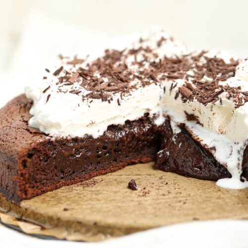A rich French molten lava cake with a soft, fudgy center oozing onto the parchment-lined board, topped with a thick layer of whipped cream and dark chocolate shavings.