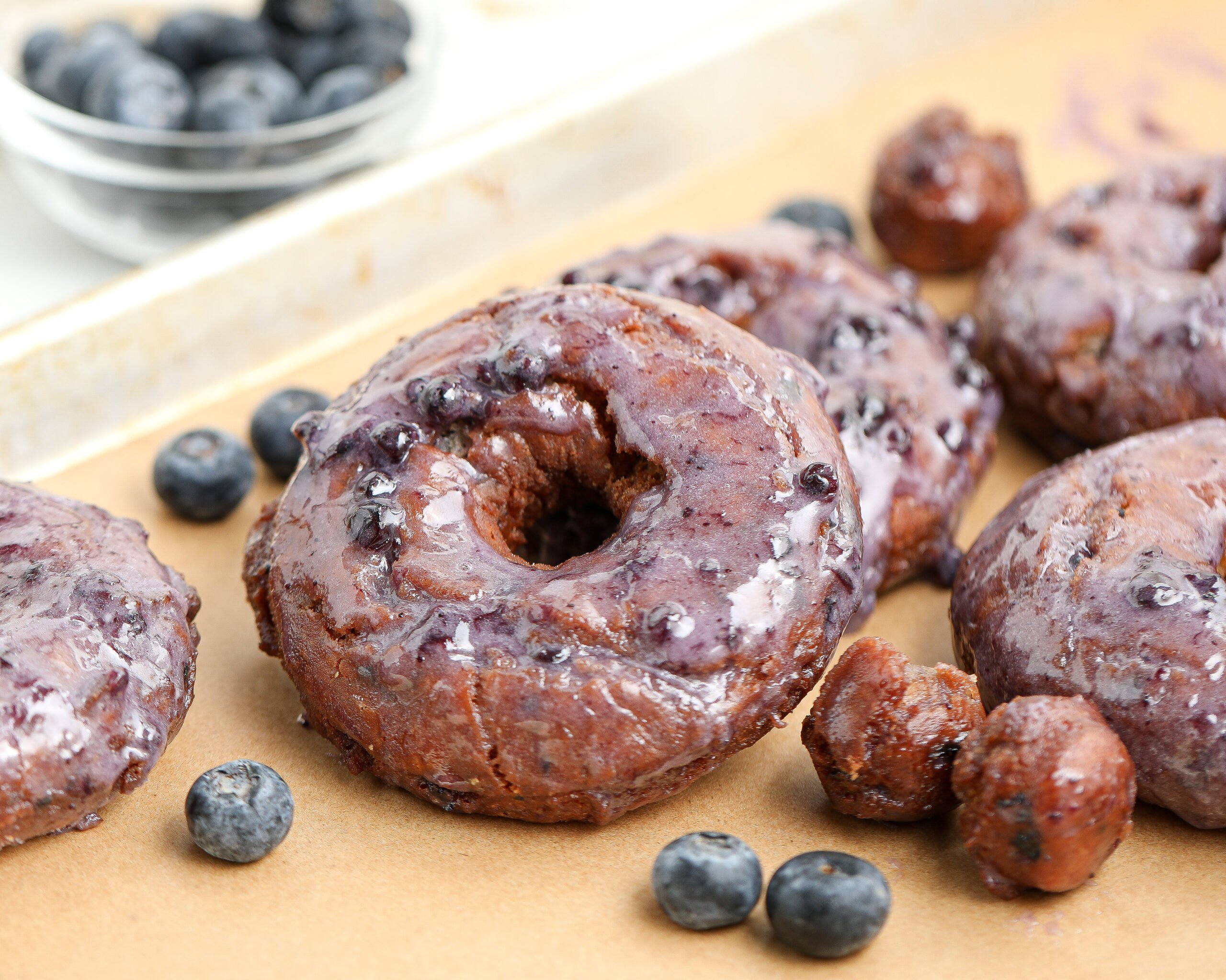 Glazed old-fashioned blueberry donuts with a glossy blueberry glaze, arranged on parchment paper with fresh blueberries scattered around.