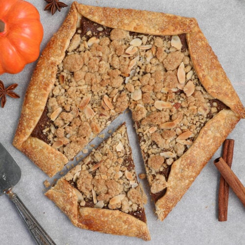 Rustic pumpkin pie galette topped with almond crumb, shown on parchment paper with a pie server, cinnamon sticks, star anise, and a mini pumpkin as decoration.