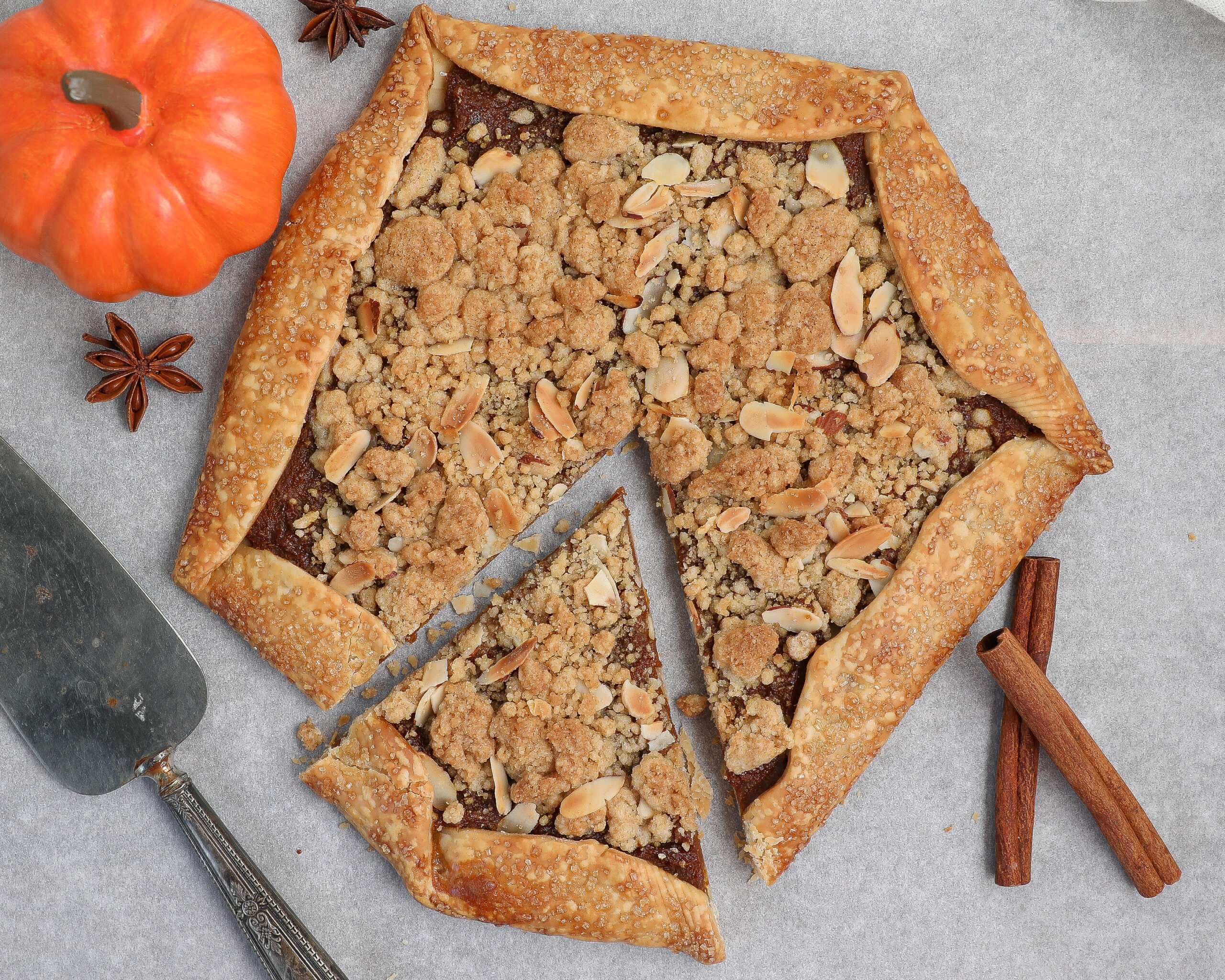 Rustic pumpkin pie galette topped with almond crumb, shown on parchment paper with a pie server, cinnamon sticks, star anise, and a mini pumpkin as decoration.