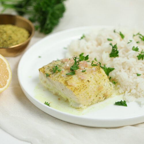 A white plate with a serving of white rice garnished with parsley and a piece of baked fish topped with sauce. In the background are a halved lemon, a bowl of seasoning, and fresh herbs.