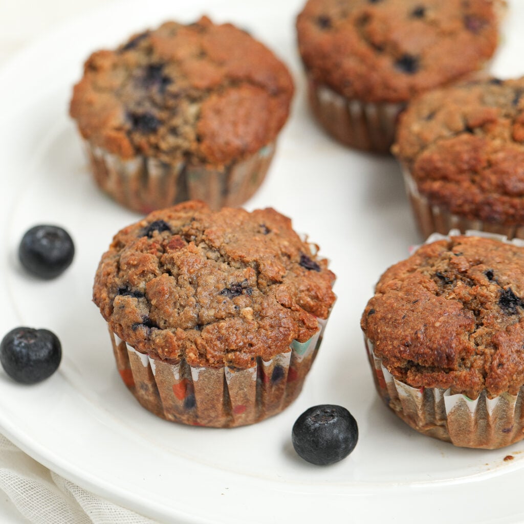 Close-up of baked blueberry oat bran muffins on a white plate, showing their golden-brown tops and visible blueberries, with a few fresh blueberries scattered around.