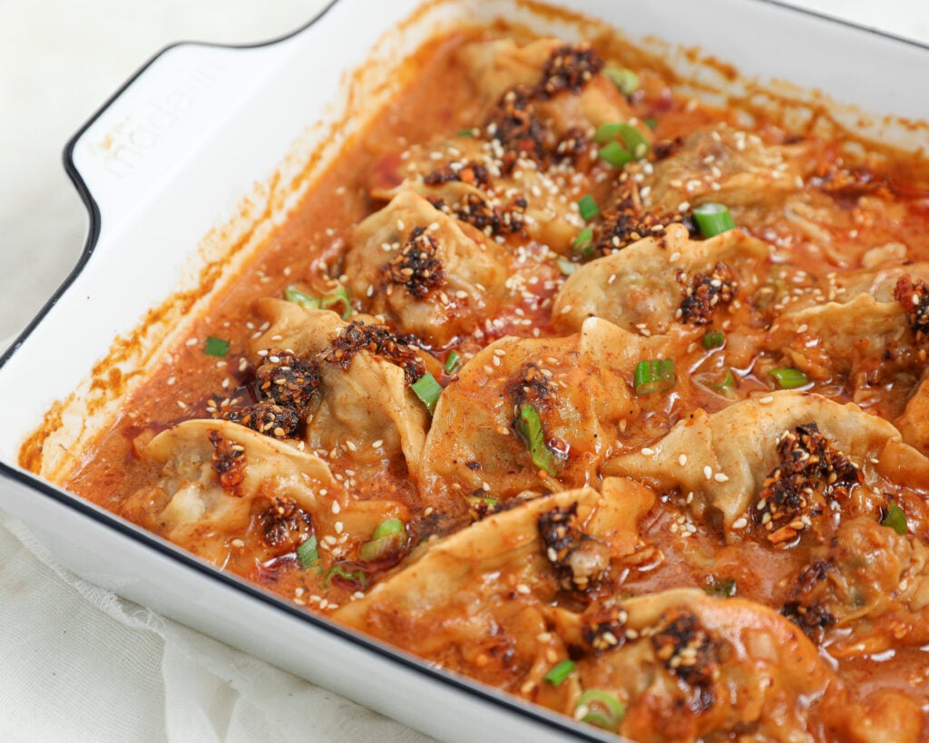 A white baking dish filled with dumplings in a rich, red sauce, garnished with sesame seeds, chopped green onions, and chili oil. The dumplings are partially submerged and arranged closely together.