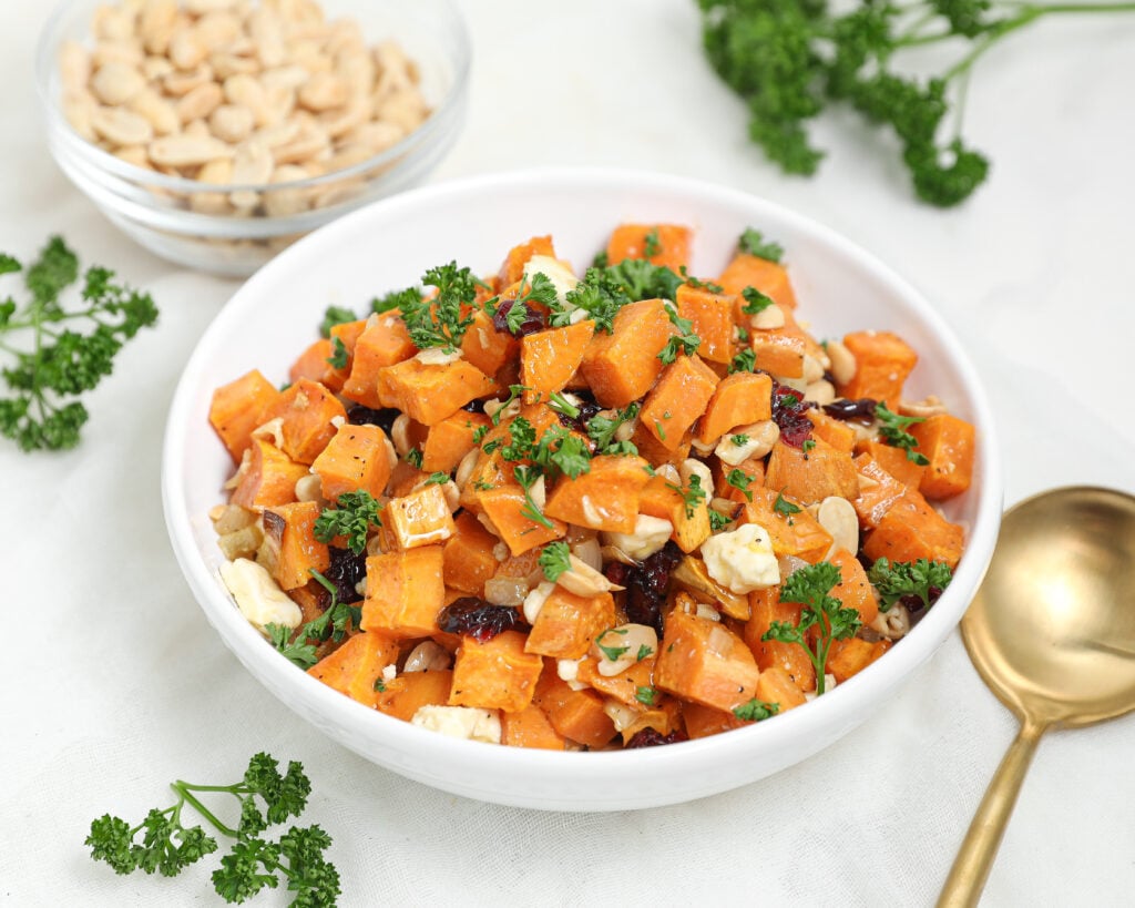 A white bowl filled with a salad of cubed roasted sweet potatoes, chopped parsley, nuts, and crumbled cheese, with a gold spoon, fresh parsley, and a bowl of nuts nearby on a light surface.
