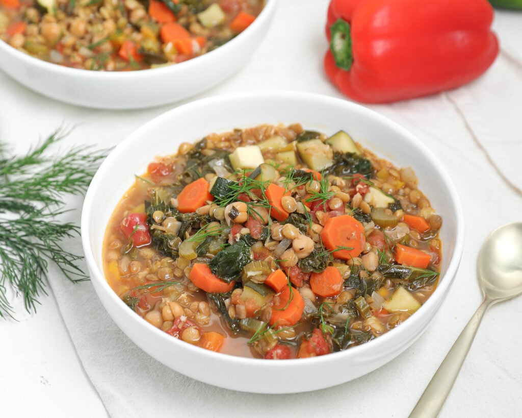 A bowl of vegetable and lentil soup with carrots, greens, and herbs, garnished with fresh dill. Another bowl of soup, a red bell pepper, a gold spoon, and sprigs of dill are nearby on a white table.