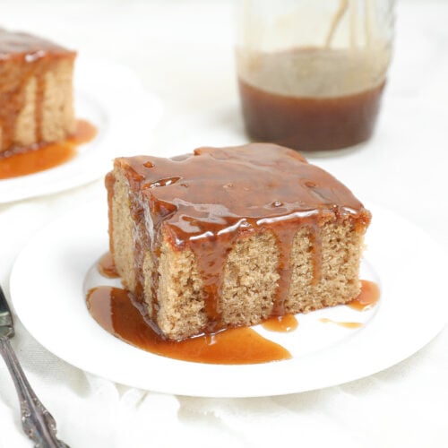 A square slice of moist biscoff sticky pudding cake covered in caramel sauce sits on a white plate, with a fork beside it and a jar of caramel sauce in the background. Another slice of cake is visible in the background.