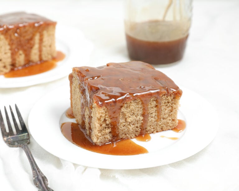 A square slice of moist biscoff sticky pudding cake covered in caramel sauce sits on a white plate, with a fork beside it and a jar of caramel sauce in the background. Another slice of cake is visible in the background.