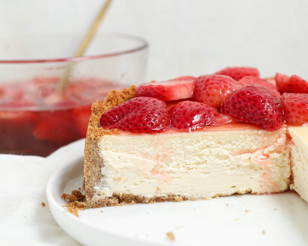 A close-up of a cheesecake with a graham cracker crust, topped with fresh strawberries. A slice is missing, showing the creamy texture inside. In the background, there is a glass bowl with more strawberries.