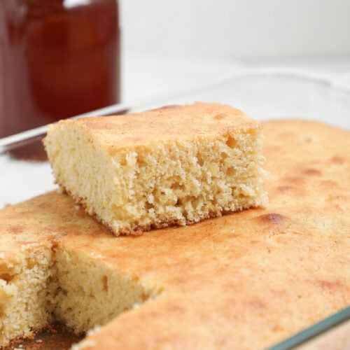 A close-up of a rectangular dish of cornbread with one square piece cut out and placed on top. A jar of honey or syrup is blurred in the background.
