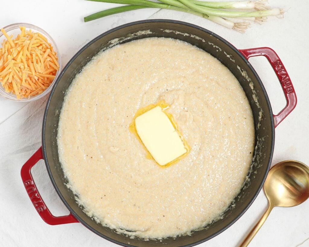 A red pot filled with creamy grits topped with a pat of melting butter. Beside the pot are a bunch of green onions, a gold ladle, and a small bowl of shredded cheddar cheese on a white surface.