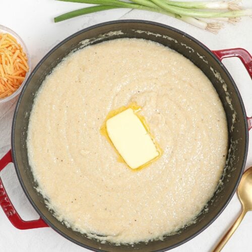 A red pot filled with creamy grits topped with a pat of melting butter. Beside the pot are a bunch of green onions, a gold ladle, and a small bowl of shredded cheddar cheese on a white surface.