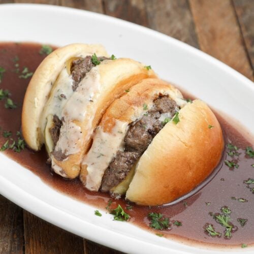 Two burger patties in a sliced bun topped with creamy sauce, served on a white oval plate with a pool of dark brown gravy and sprinkled with chopped herbs. The plate rests on a rustic wooden surface.