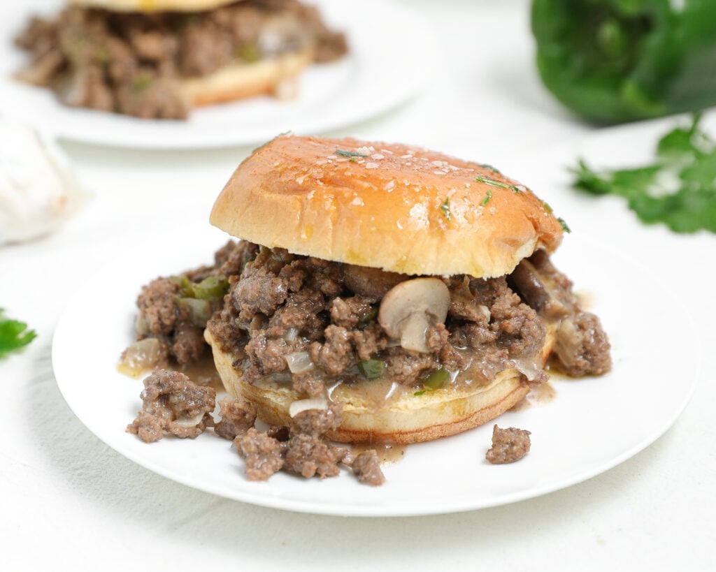 A sandwich filled with cooked ground beef, mushrooms, and diced green peppers sits on a white plate. The filling is spilling out from between a golden toasted bun. Another sandwich is blurred in the background.