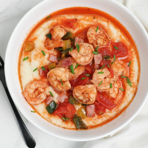 A white bowl filled with creamy grits topped with shrimp, diced tomatoes, green peppers, and herbs, set on a white surface next to a black fork.