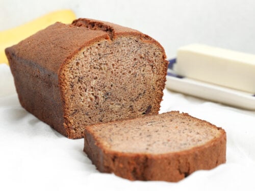 A loaf of banana bread with one slice cut and placed in front, showing a moist, tender crumb and golden brown crust on a white surface.