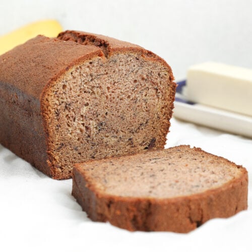 A loaf of banana bread with one slice cut and placed in front, showing a moist, tender crumb and golden brown crust on a white surface.