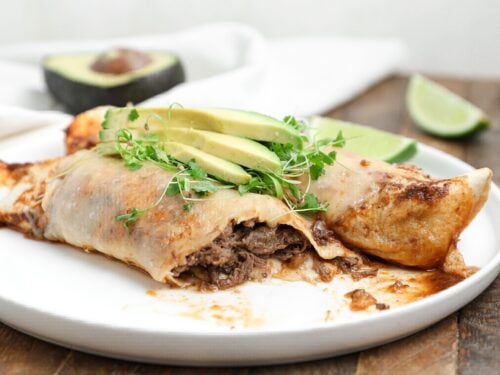 Plate of beef barbacoa enchiladas topped with avocado slices and microgreens, with lime wedges and half an avocado in the background.