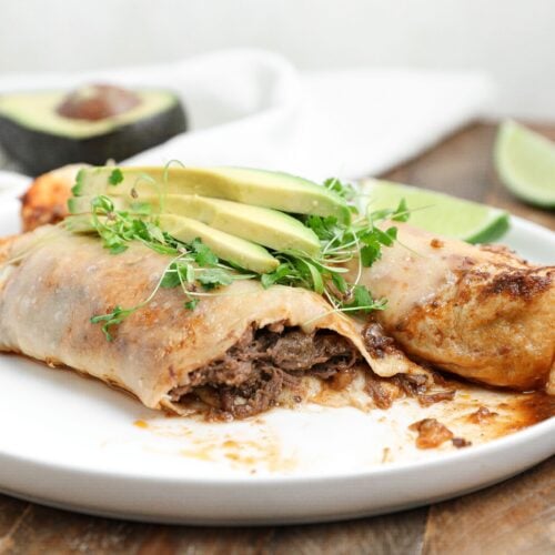 Plate of beef barbacoa enchiladas topped with avocado slices and microgreens, with lime wedges and half an avocado in the background.