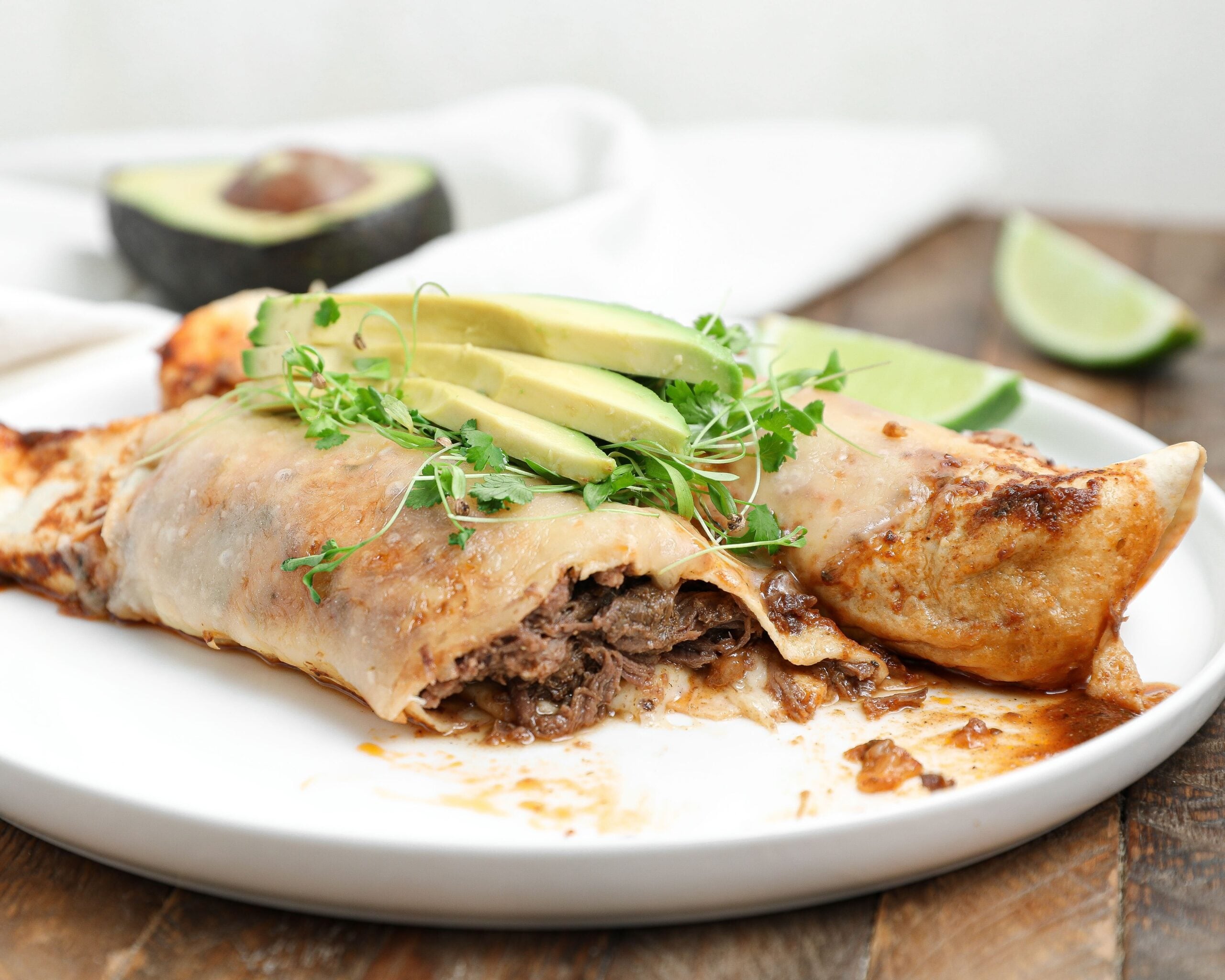 Plate of beef barbacoa enchiladas topped with avocado slices and microgreens, with lime wedges and half an avocado in the background.