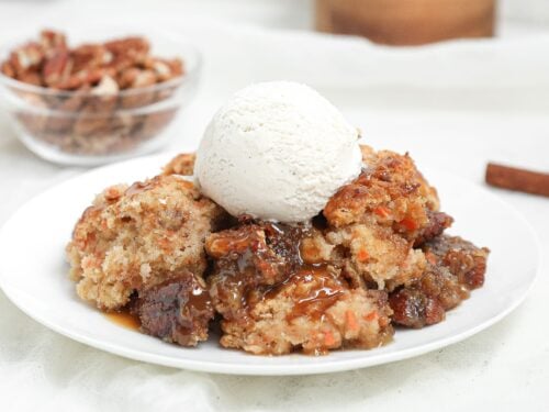 Warm carrot cake cobbler with gooey caramel pecan filling topped with a scoop of vanilla ice cream on a white plate.