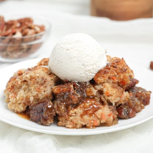 Warm carrot cake cobbler with gooey caramel pecan filling topped with a scoop of vanilla ice cream on a white plate.