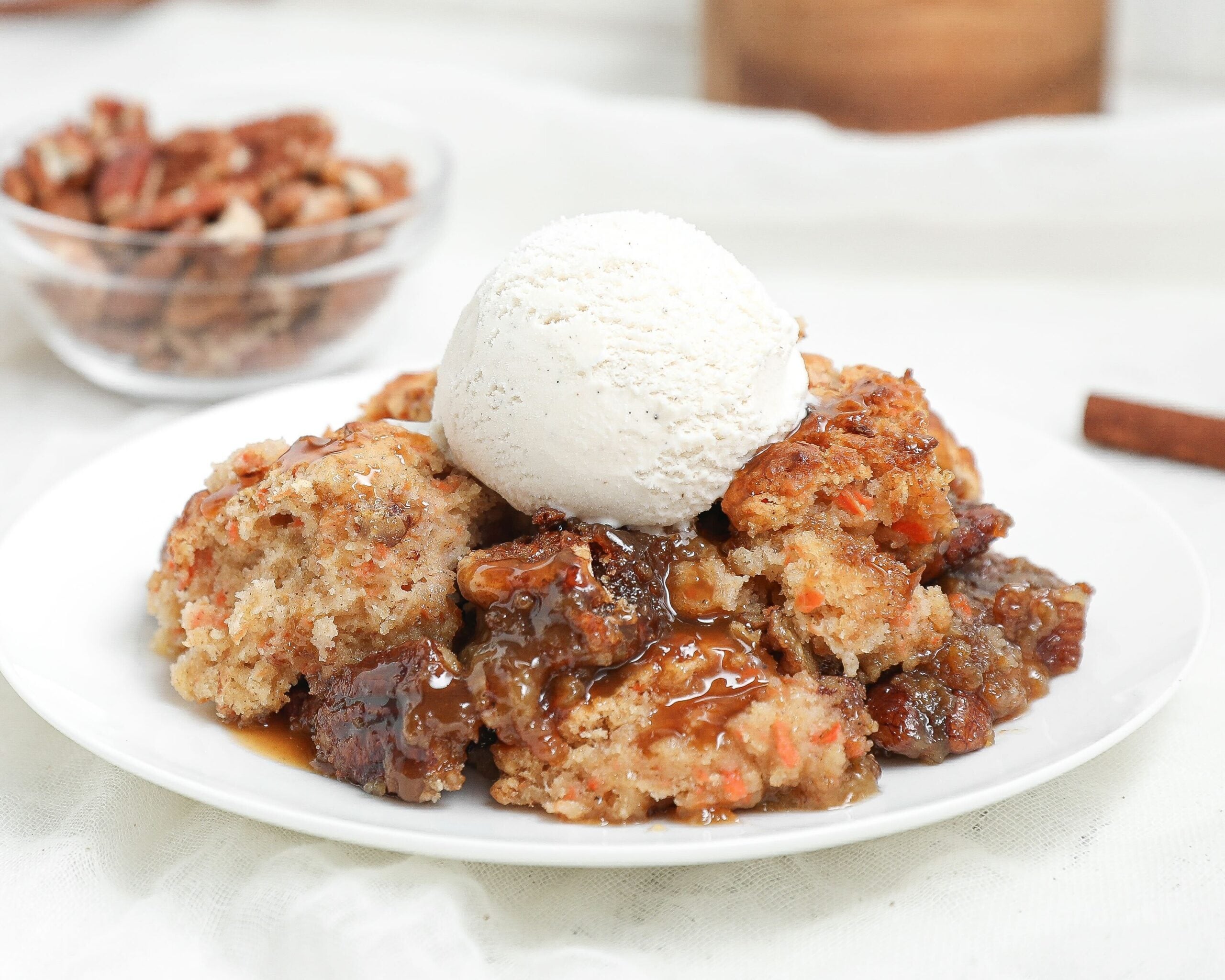 Warm carrot cake cobbler with gooey caramel pecan filling topped with a scoop of vanilla ice cream on a white plate.