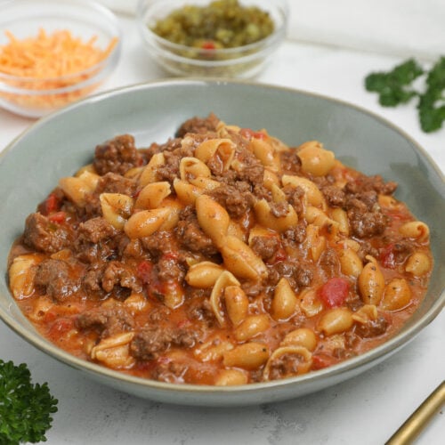 Bowl of cheeseburger pasta with shell pasta, ground beef, and a cheesy tomato-based sauce, with shredded cheddar and relish blurred in the background.