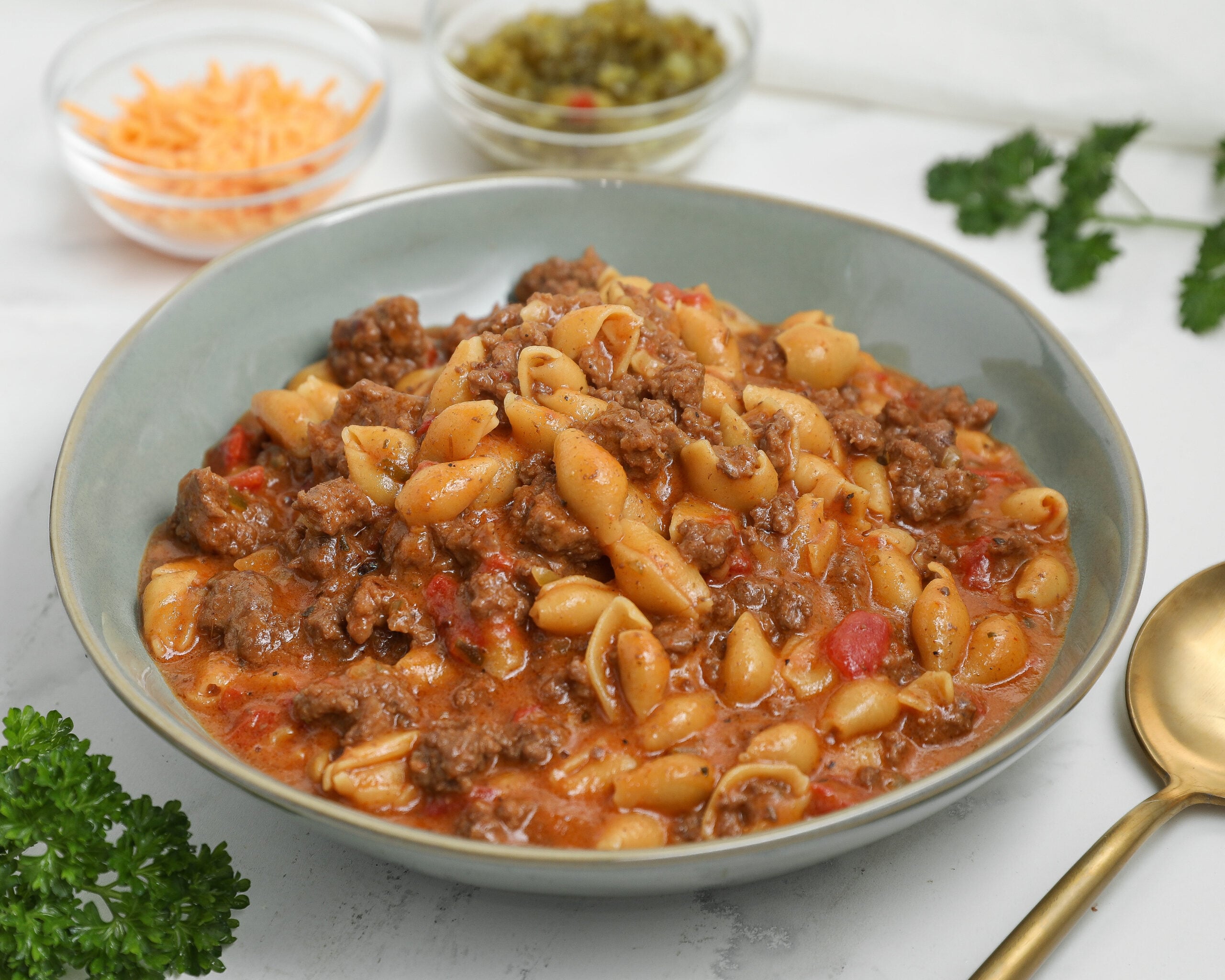 Bowl of cheeseburger pasta with shell pasta, ground beef, and a cheesy tomato-based sauce, with shredded cheddar and relish blurred in the background.