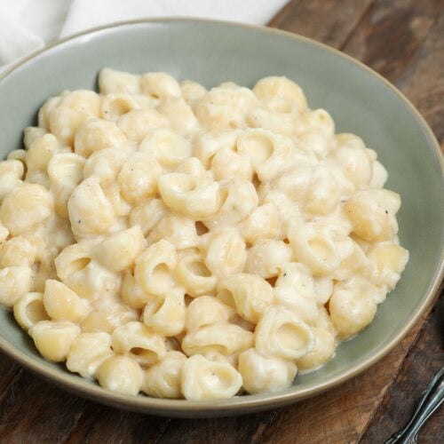 Bowl of creamy white cheddar mac and cheese with shell pasta on a wooden table, with a spoon beside the bowl.
