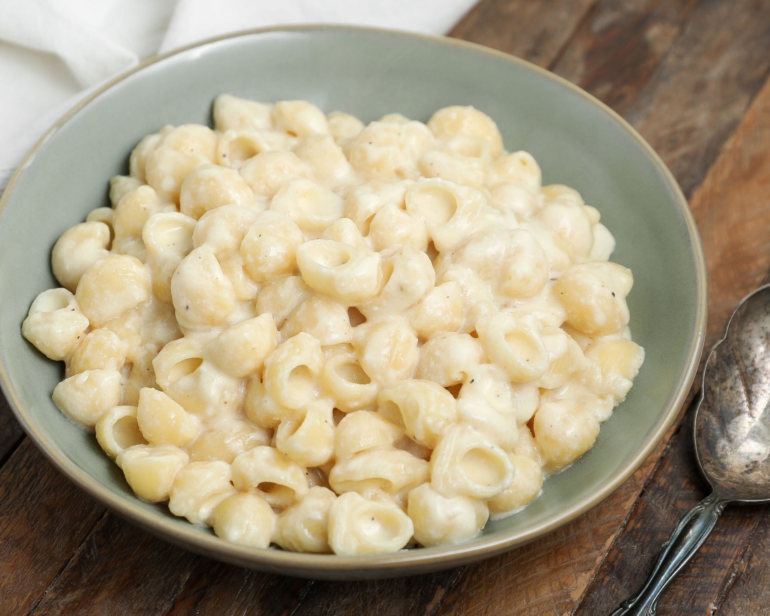 Bowl of creamy white cheddar mac and cheese with shell pasta on a wooden table, with a spoon beside the bowl.